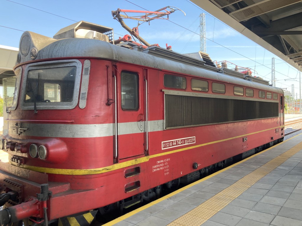 A red electric train at a station, featuring a classic design with large windows and details on the side, set against a clear blue sky.