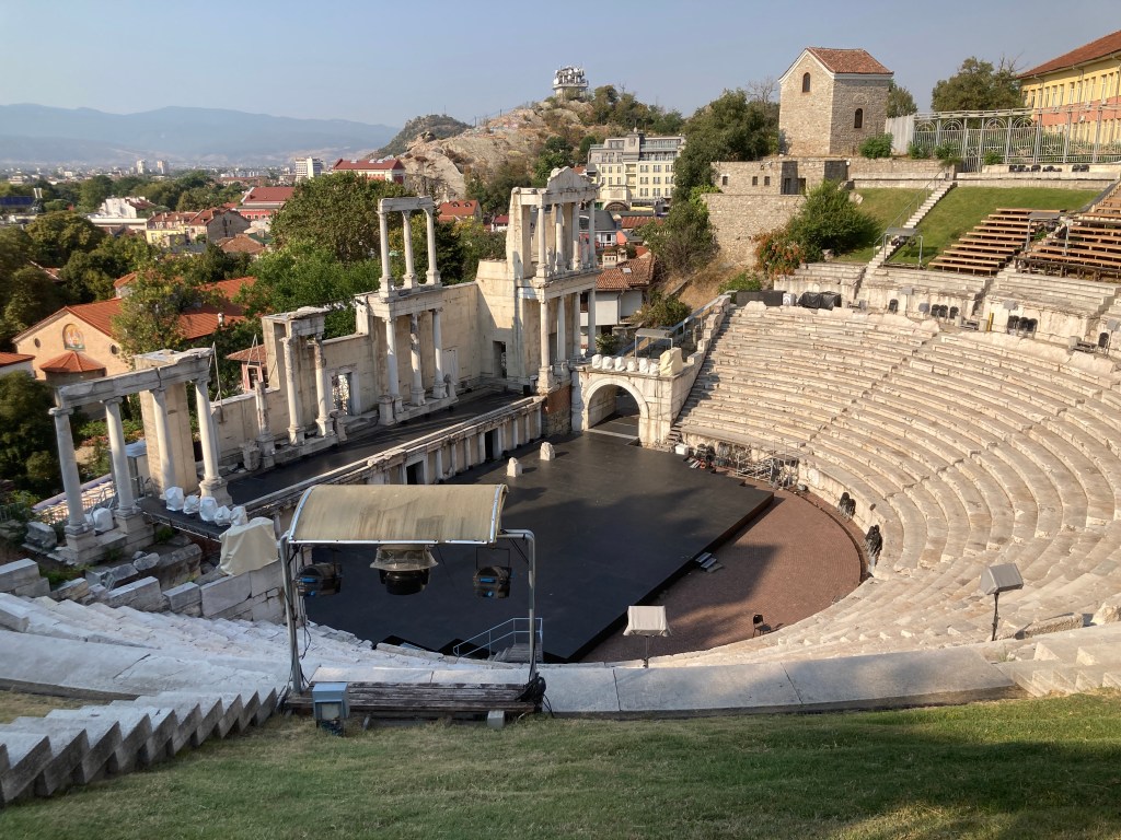 A panoramic view of an ancient Roman amphitheatre featuring stone seating and partially preserved columns, set against a backdrop of hills and modern buildings.