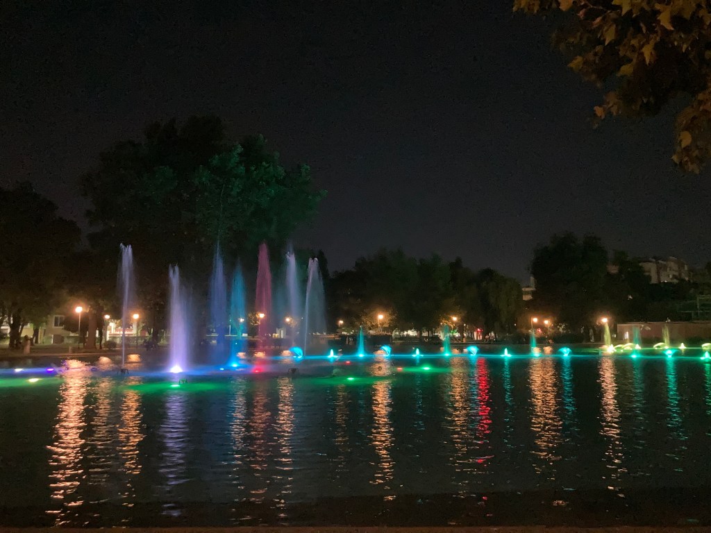 A beautifully illuminated fountain at night, with colourful lights reflecting on the water surface, surrounded by trees and faintly lit park pathways.