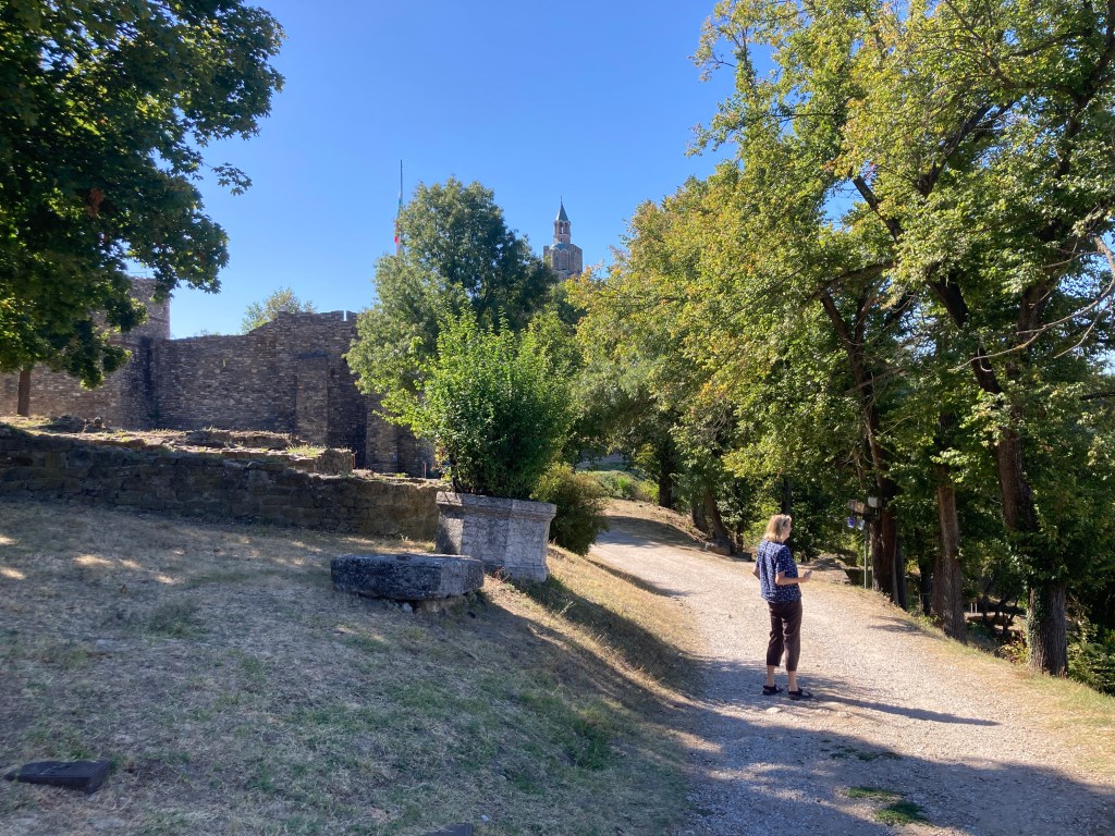 A person standing on a gravel path near the ruins of an old stone structure, surrounded by lush trees and greenery, with a tower visible in the background under a clear blue sky.